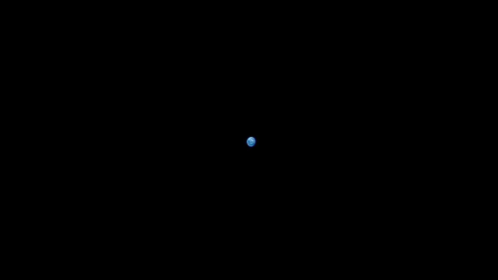La Tierra como una pequeña bola blanco azulada en medio del negro del espacio vista desde las una de las cámaras de la cápsula Orión de la misión