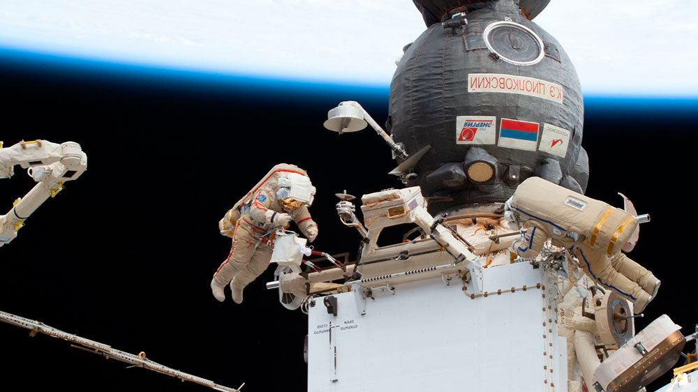 Los dos cosmonautas trabajando en el radiador con una cápsula Soyuz y la Tierra al fondo