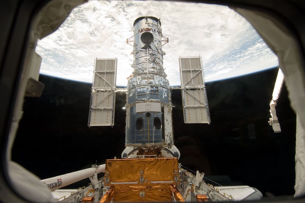 La bodega de carga del Atlantis fotografiada a través de una de sus ventanas. Al fondo de ella está el Hubble en posición vertical, convenientemente sujeto. La Tierra hace de fondo de la foto