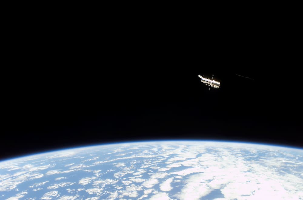 El Hubble flotando en la negrura del espacio con la Tierra ocupando la mitad inferior de la foto. El telescopio se ve a propósito pequeño en la imagen para dar una ligera idea de escala