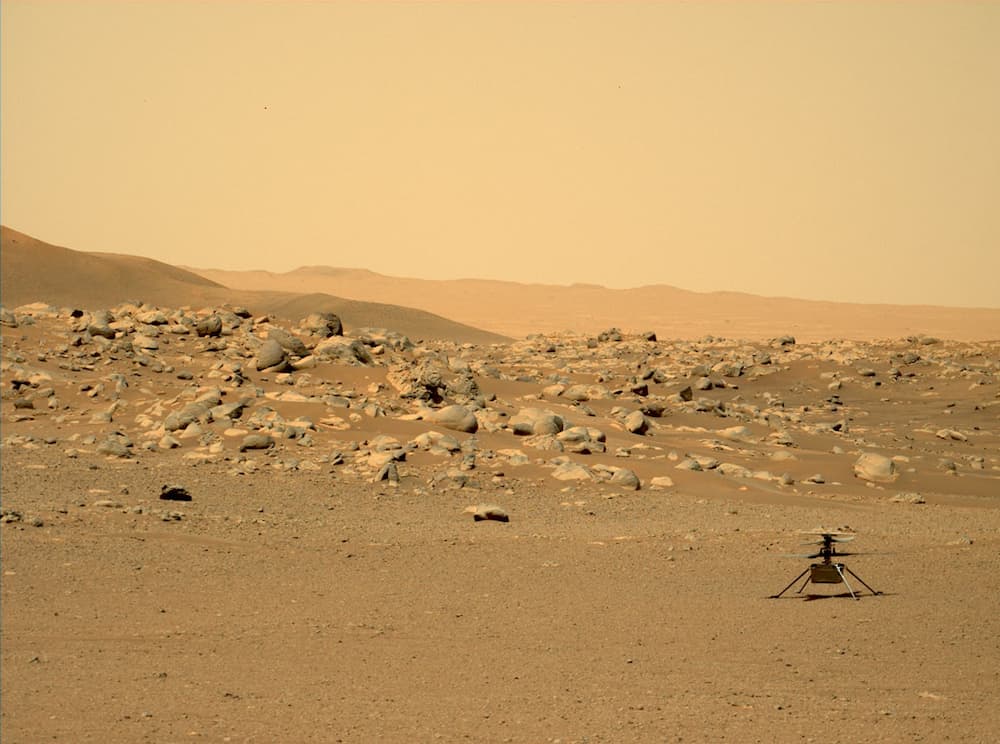 Ingenuity en una zona plana de la superficie de Marte con rocas y elevaciones al fondo