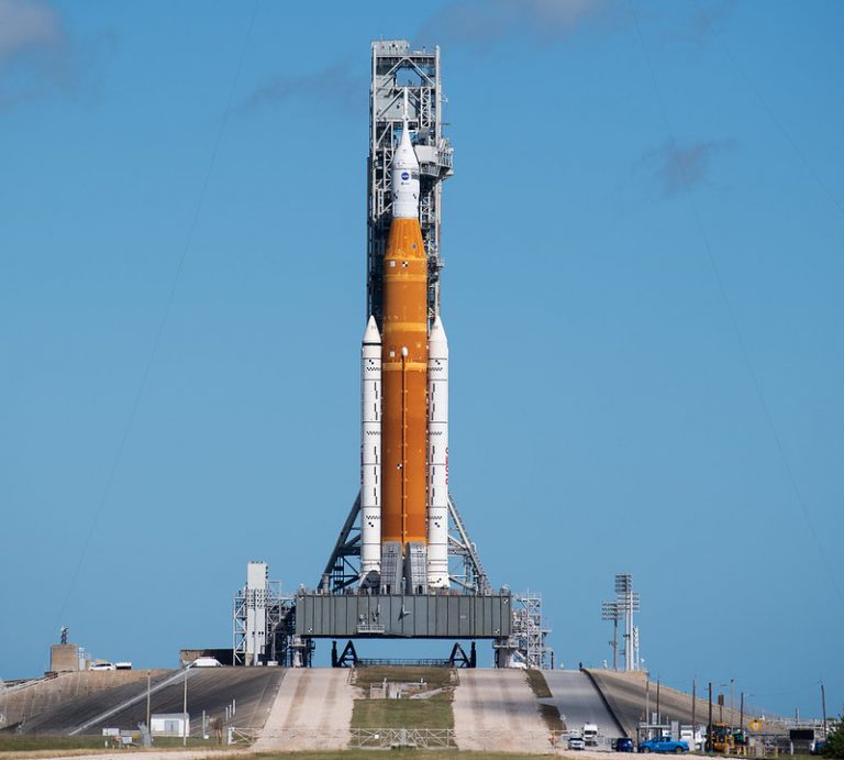 El SLS visto de frente sobre su transporte en lo alto de la plataforma de lanzamiento con un cielo azul de fondo en el que apenas queda alguna nube