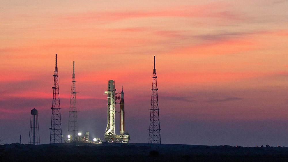 El SLS en la plataforma de lanzamiento al amanecer, iluminado por focos. De fondo, un cielo con algunas nubes con tono rojizo