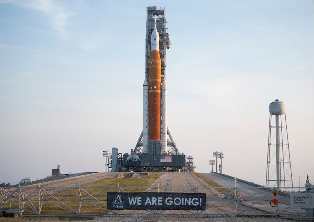 El SLS y su torre de lanzamiento sobre la plataforma de lanzamiento 39B vistos desde fuera de la valla que cierra el recinto y que tiene una pancarta que dice «We Are Going» – NASA/Joel Kowsky