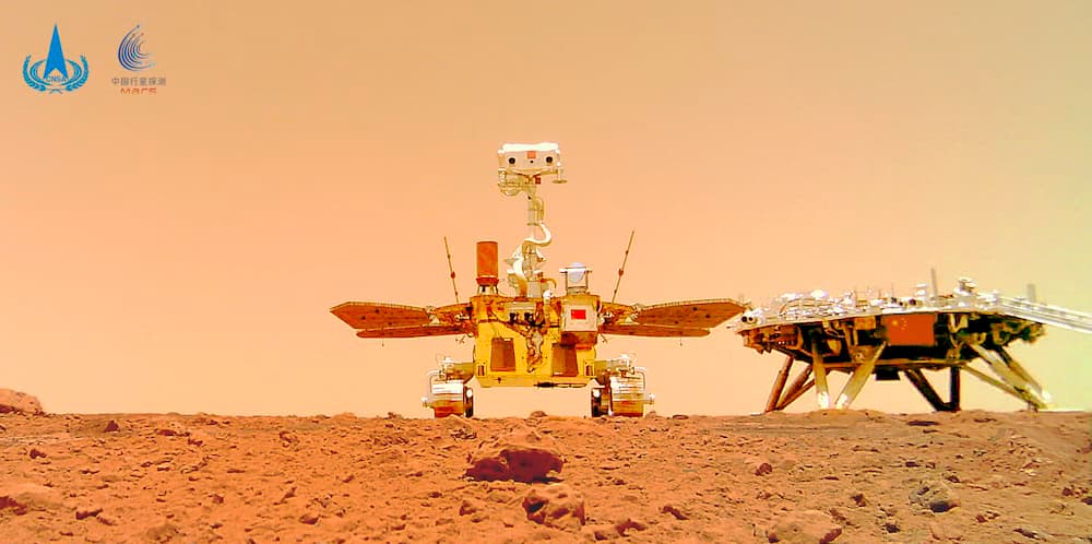 El rover Zhurong sobre la superficie de Marte en primer plano y más atrás y a la derecha su plataforma de aterrizaje – CNSA/PEC