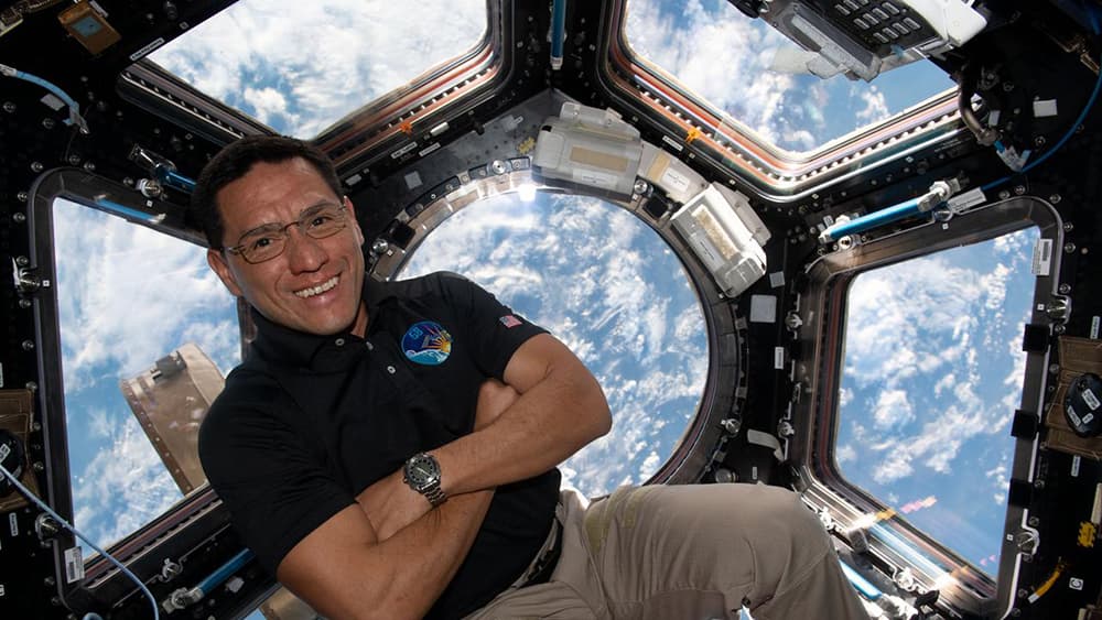Frank Rubio flota en la cúpula de la estación espacial. Tiene los brazos cruzados y sonríe a la cámara. La Tierra está al fondo, mostrando sus nubes dispersas.
