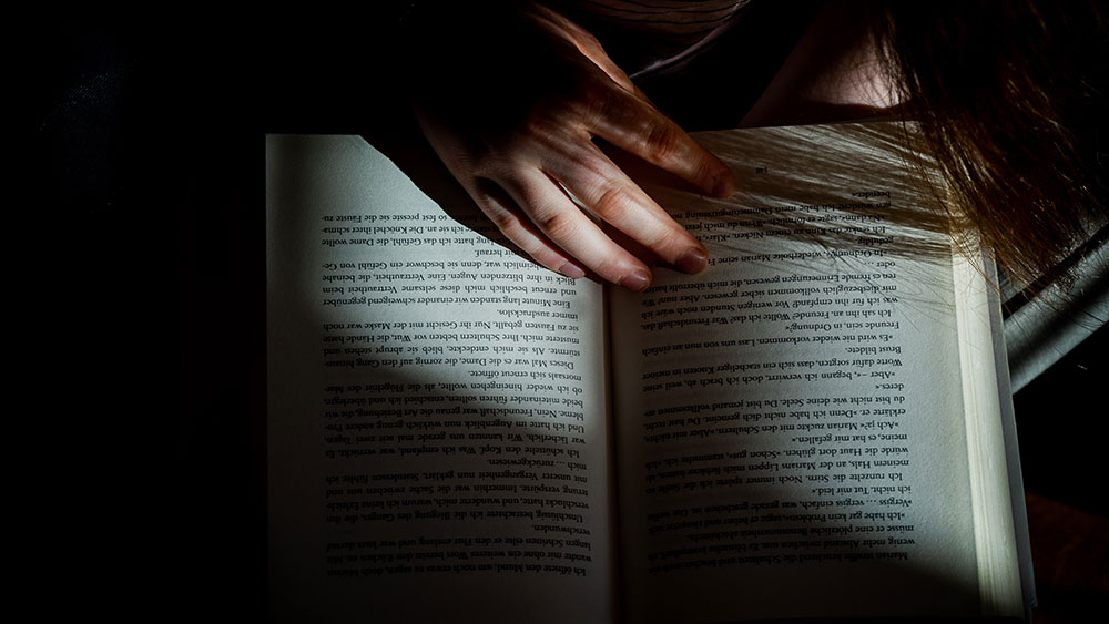 Foto cenital de un libro abierto en la sombra sobre el que cae un rayo de luz y en el que está apoyada la mano de una chica que suponemos que lo está leyendo
