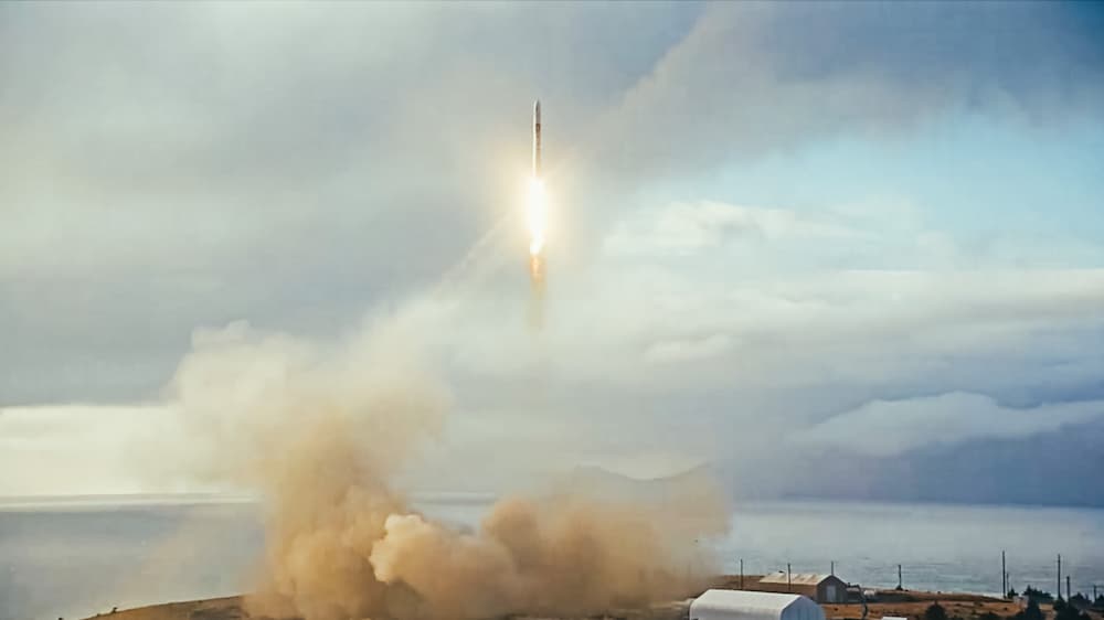 El cohete elevándose sobre la plataforma de lanzamiento con el mar y un cielo nublado de fondo