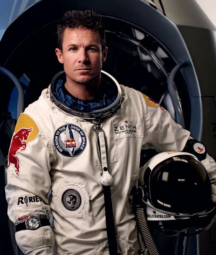 Retrato de Baumgartner con el traje que utilizó para su salto desde la estratosfera en el que además sujeta el casco bajo su brazo izquierdo; al fondo está la cápsula en la que realizó el ascenso 