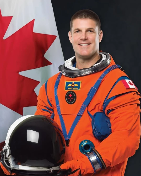 Retrato oficial de Hansen con su traje intravehicular de la misión Artemisa II; hay una bandera de Canadá al fondo