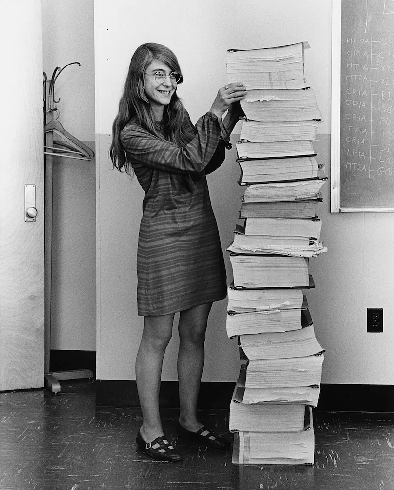 Margaret Hamilton de pie al lado de una pila de doce tomos con el softare impreso que es tan alta como ella